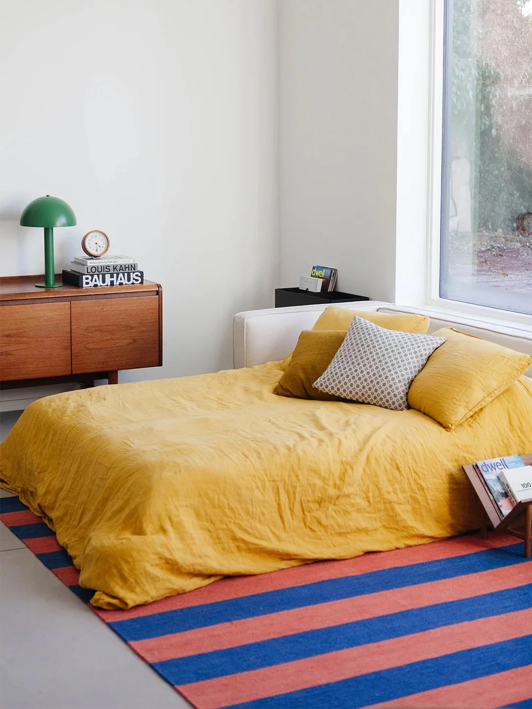 Yellow bedroom bedding lies beside a wood cabinet and green dome-shaped lamp above a blue and coral striped rug near a large window