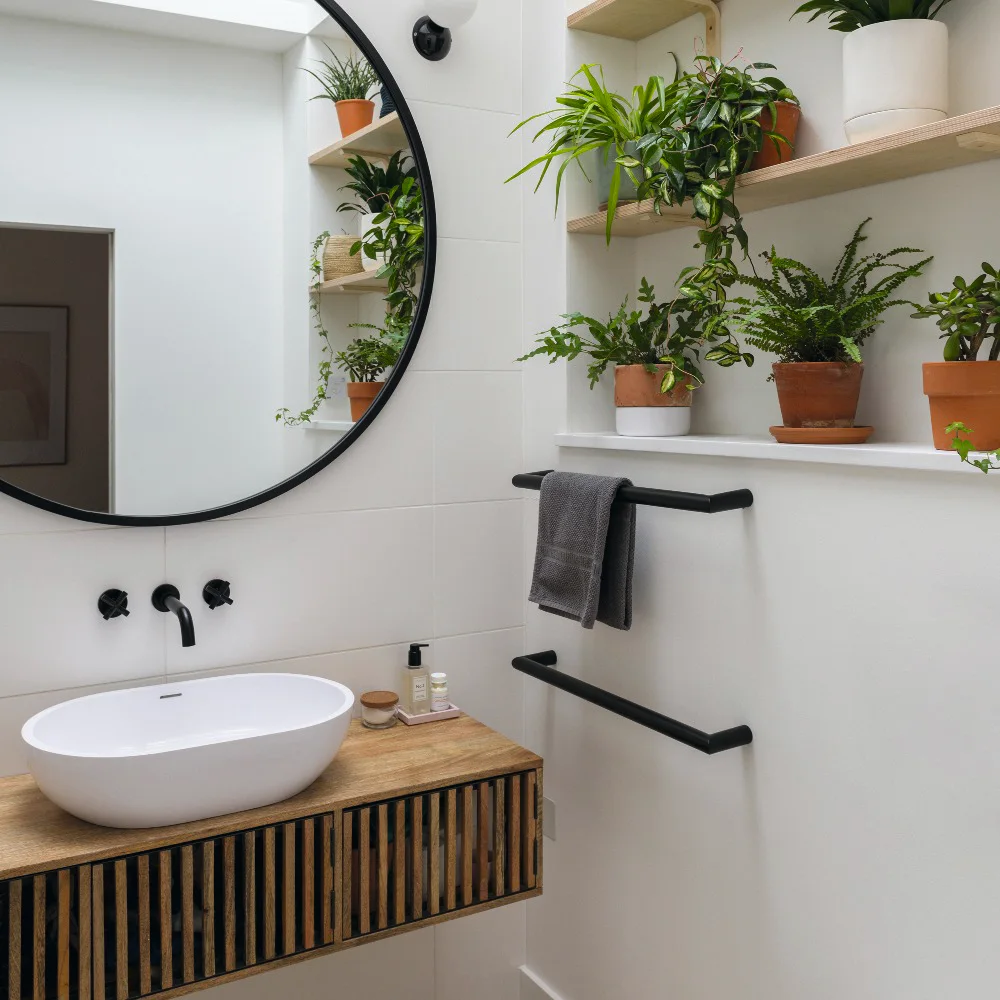 A wood-fronted floating vanity with a white basin sits below a dark round mirror beside open shelves holding plants and black towel rails