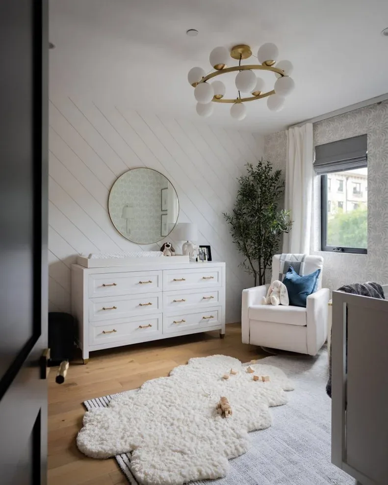 A white nursery dresser and pale armchair sit beside a diagonal white board wall, round mirror, dark window shade and layered pale rugs