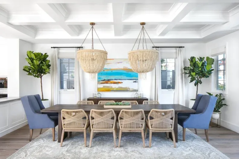 Dining room with dark rectangular table, blue upholstered end chairs, pale woven side chairs, paired fringed lights and coastal artwork.
