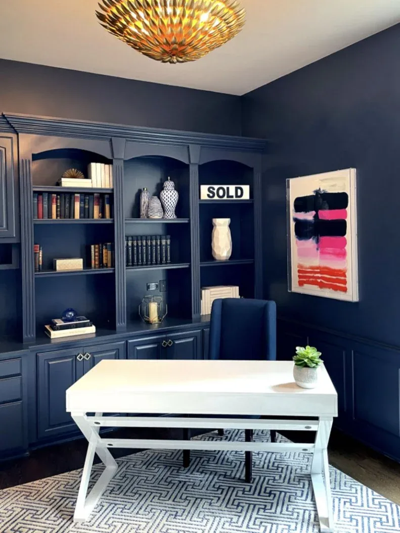 White desk in a navy office with built-in shelves and a gold-toned sculptural ceiling light.