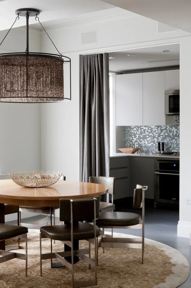 Dark low-backed dining chairs with slim frames around a round wooden table, near gray curtains and an adjoining kitchen.