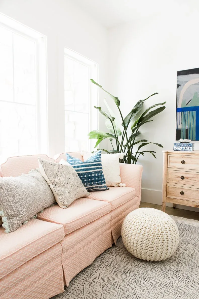 Bright living room with pale pink scalloped-back sofa, patterned light cushions, blue striped pillow, knitted pouf, wooden drawer chest and tall plant.
