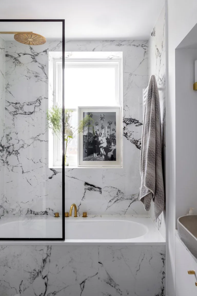 A white bathtub has strongly veined black-and-white wall and bath-panel surfaces beside a black screen edge and gold-colored taps