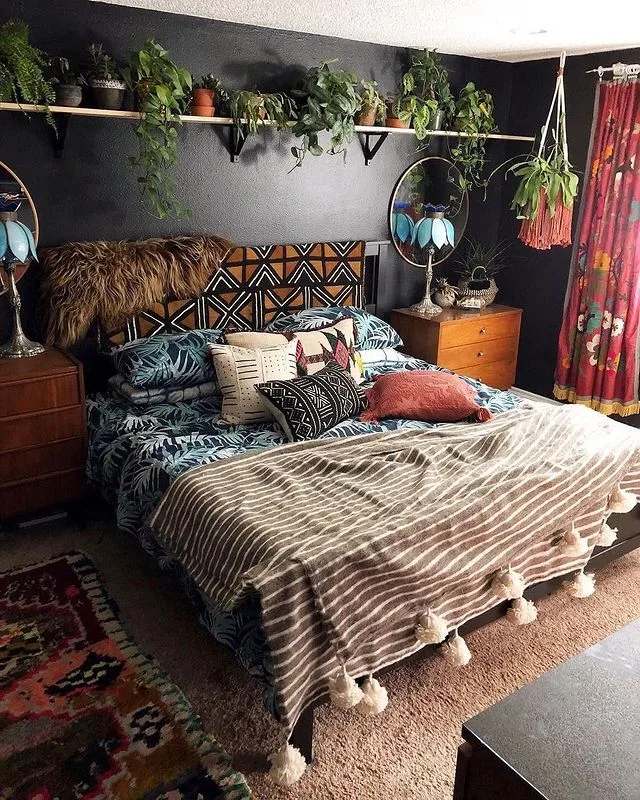 Black bedroom walls with a plant shelf, patterned headboard and bedding, tasseled striped throw, and red printed curtains.