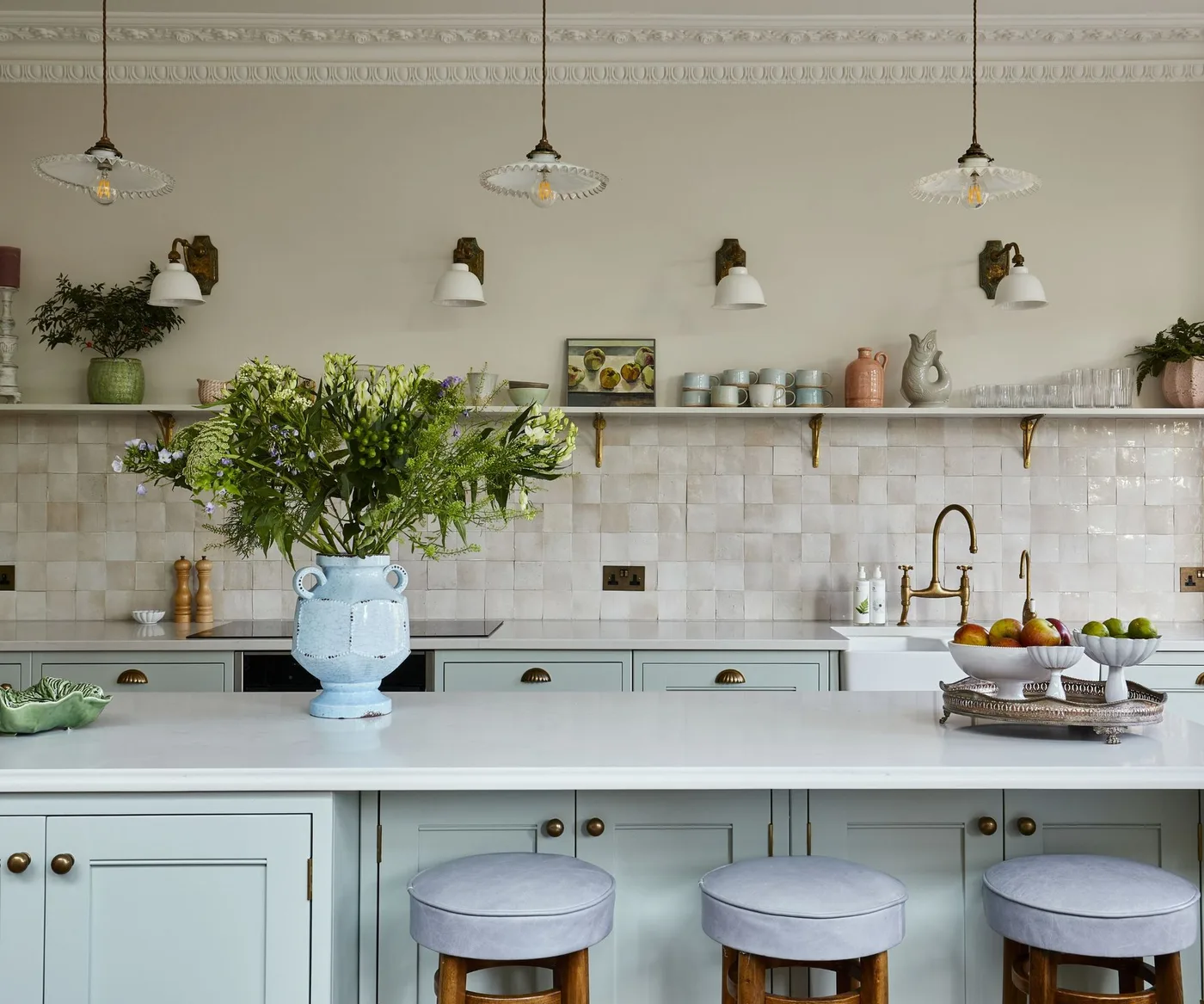 Pale blue kitchen cabinets with glossy cream square tiles in an offset pattern, a long shelf and three small wall lights.