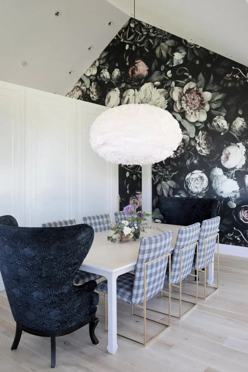 Dining room with large pale flowers on a black feature wall, white paneled side wall, white table, checked chairs, dark upholstered end chairs, and pale pendant.