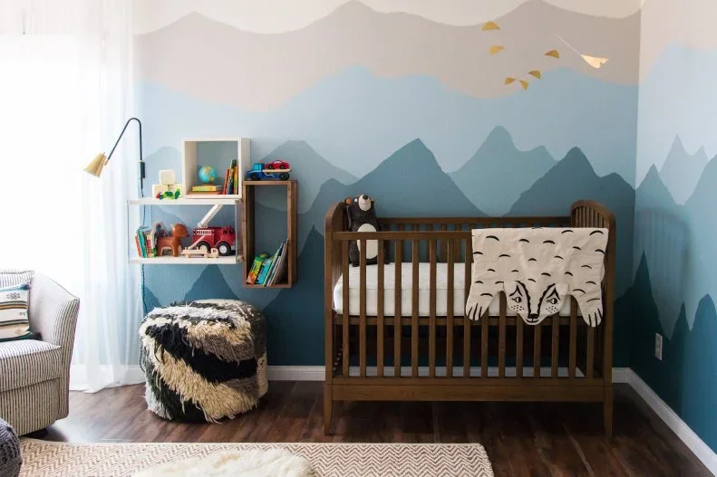 A wood crib stands beside a blue layered mountain mural, small open book boxes, a pale chair and a dark patterned pouf in a nursery