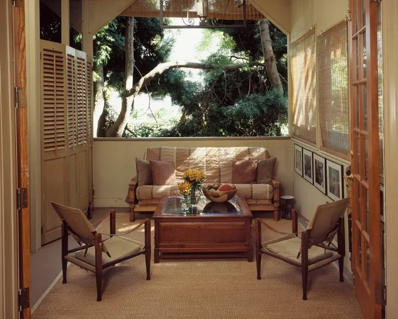 Sheltered open-air parlor with wooden chairs, pale woven sofa, warm wooden coffee table, light floor covering and garden view.