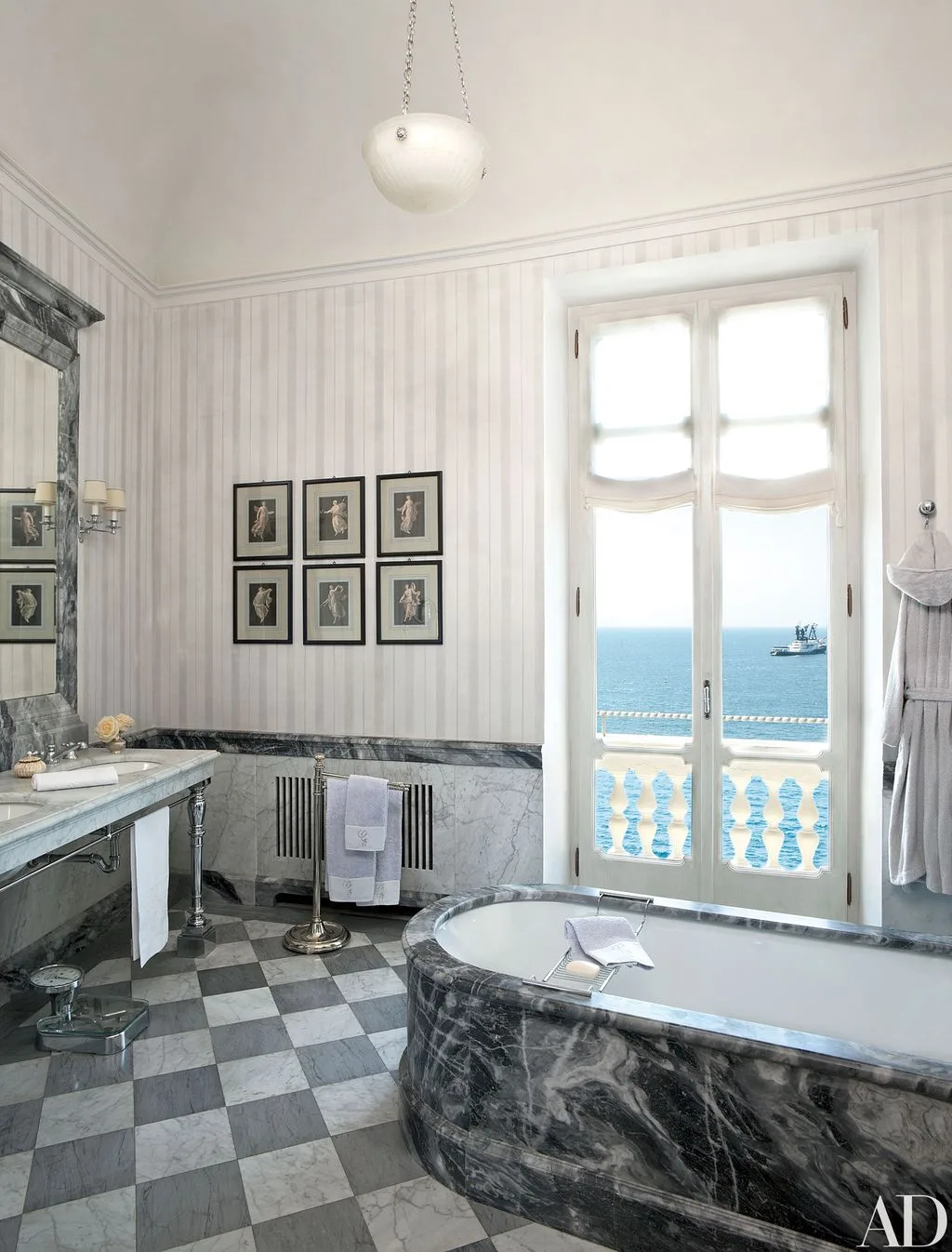 Bathroom with a deep gray veined tub surround, gray checkerboard floor, pale striped walls, framed pictures, and bright French doors.