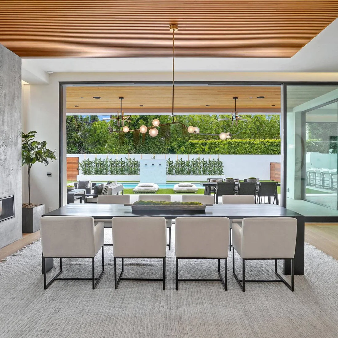 A dark rectangular dining table and pale upholstered chairs sit on a gray rug under a slatted wood ceiling beside open glass doors and outdoor seating