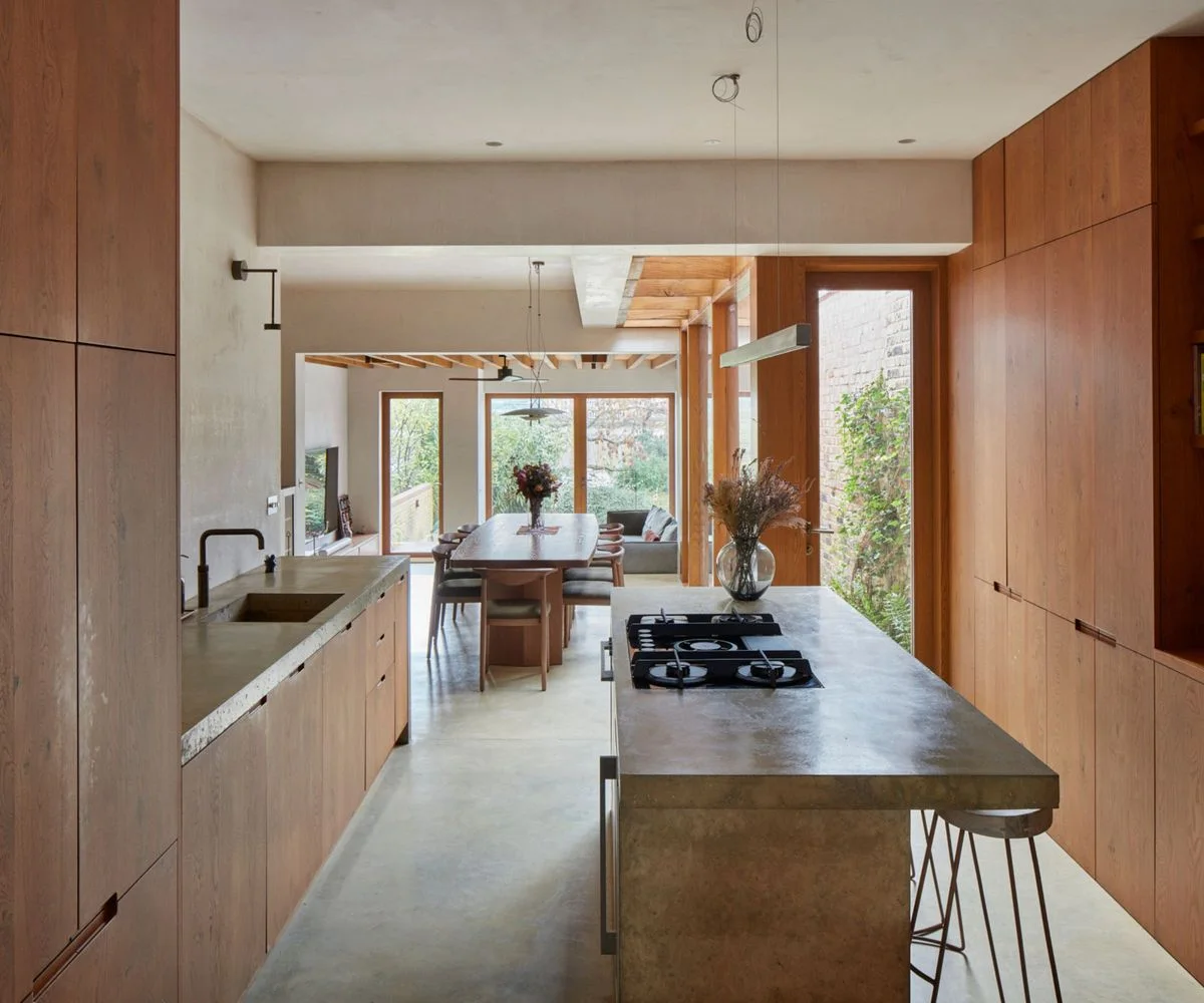 Wooden storage walls surround a narrow gray-topped island and sink counter beside a dining table, wooden ceiling details and garden-facing windows
