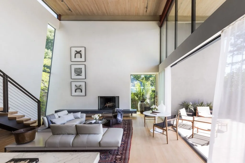 A small dark rectangular fireplace opening sits low in a tall white wall beside a staircase, pale sectional seating, broad glazing and three small framed artworks