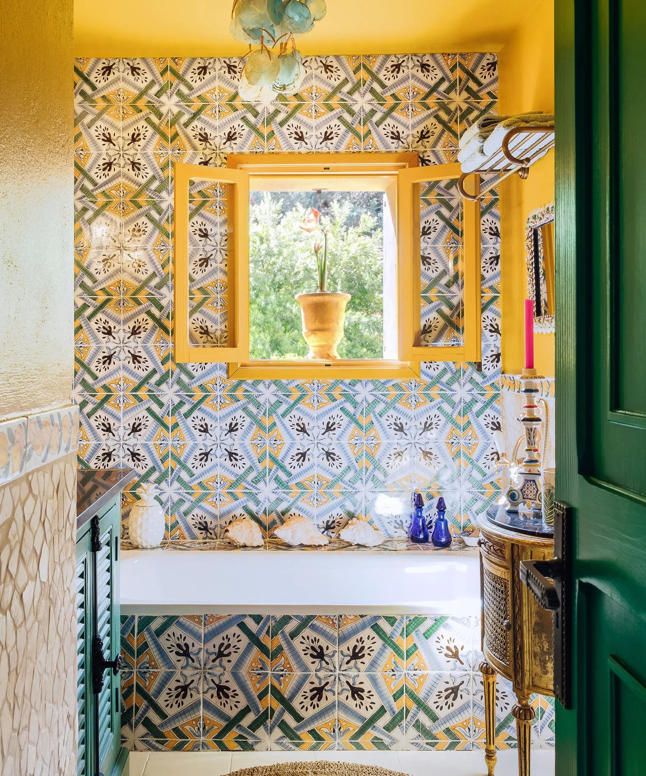 Yellow paint frames a window above a bath surrounded by multicolored patterned tiles, decorative vessels and a green door