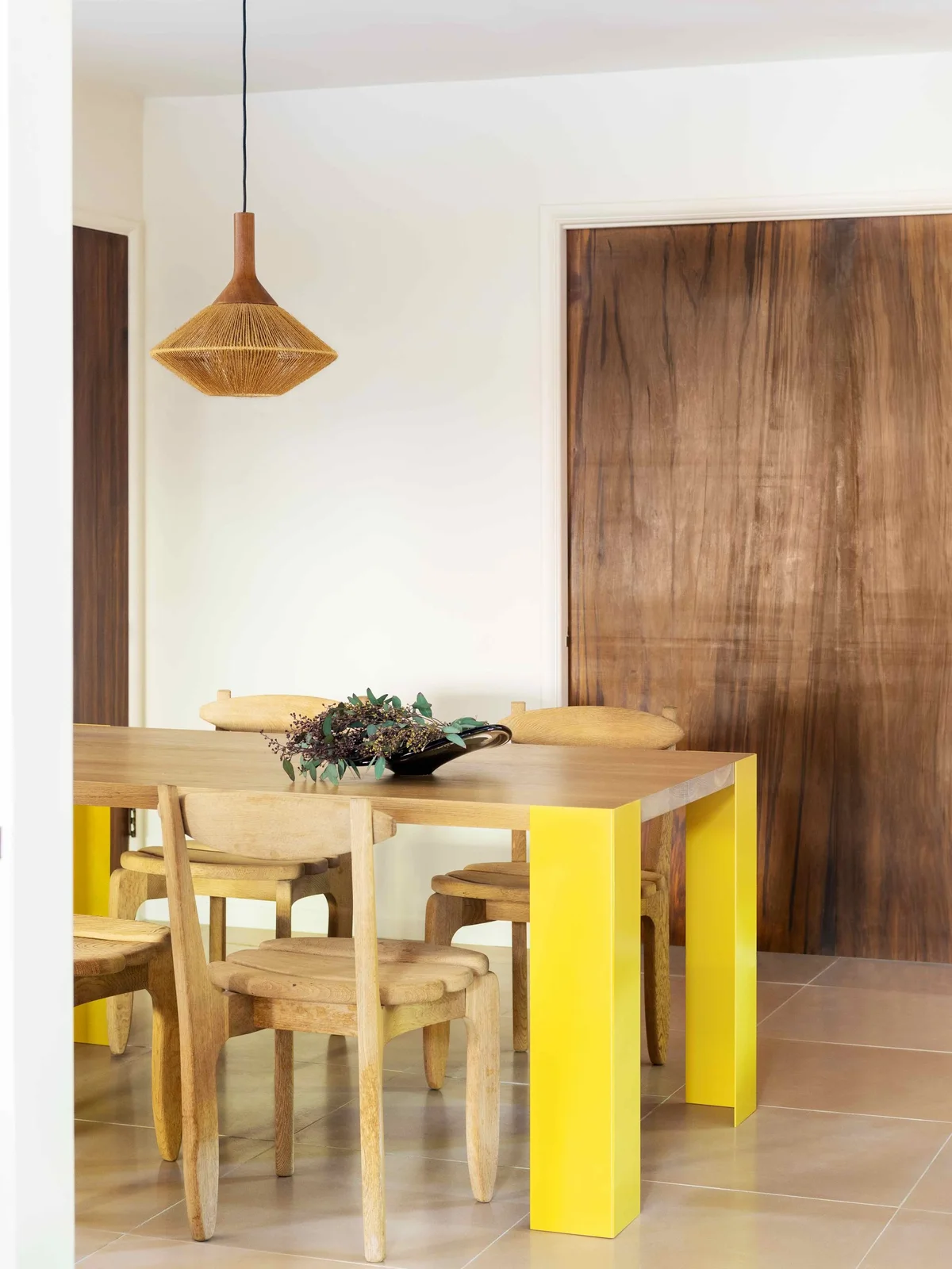 A wood dining tabletop stands on bright yellow supports among curved pale wood chairs beneath a woven pendant beside white walls and a broad wood door