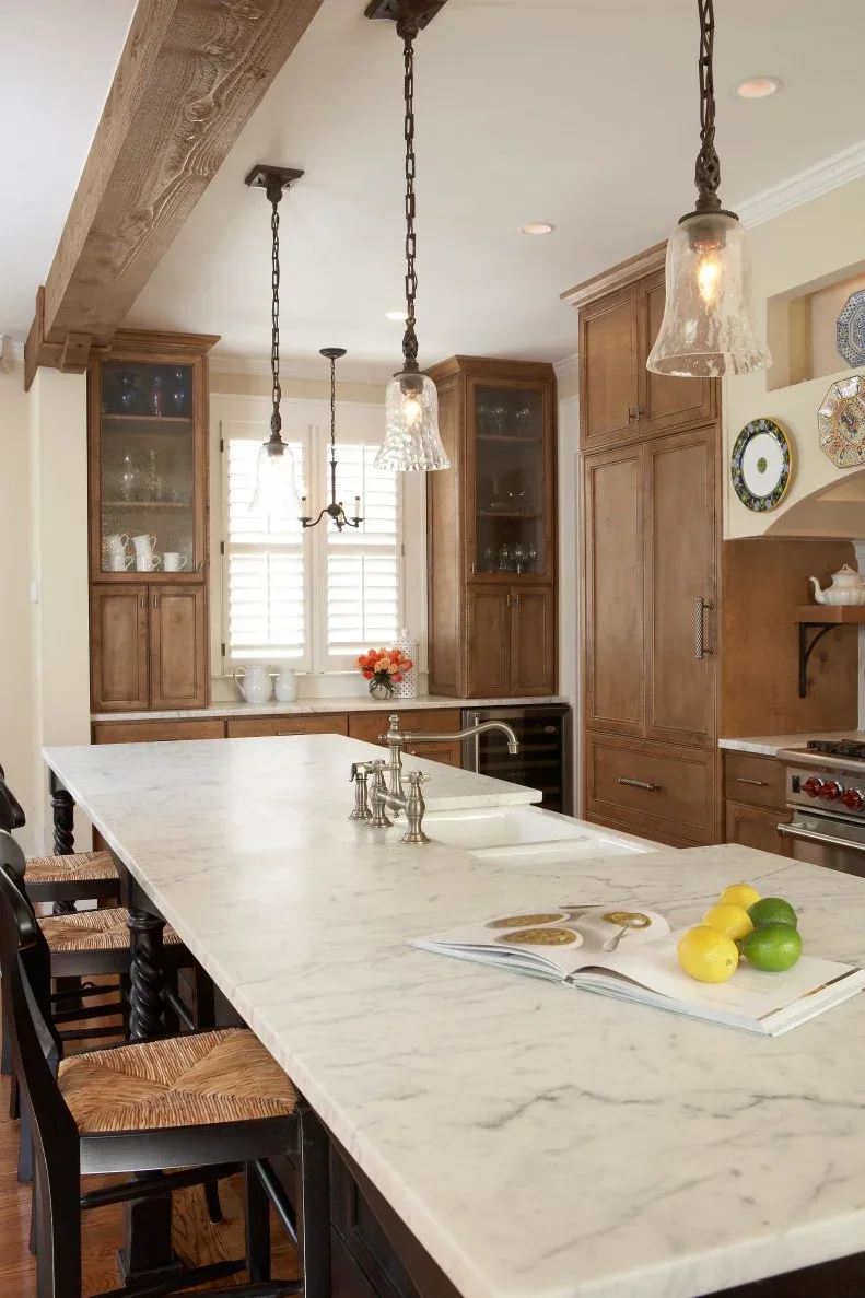 Traditional wood kitchen cabinets with pale island, glass-front upper cupboards, hanging glass lights and an exposed beam