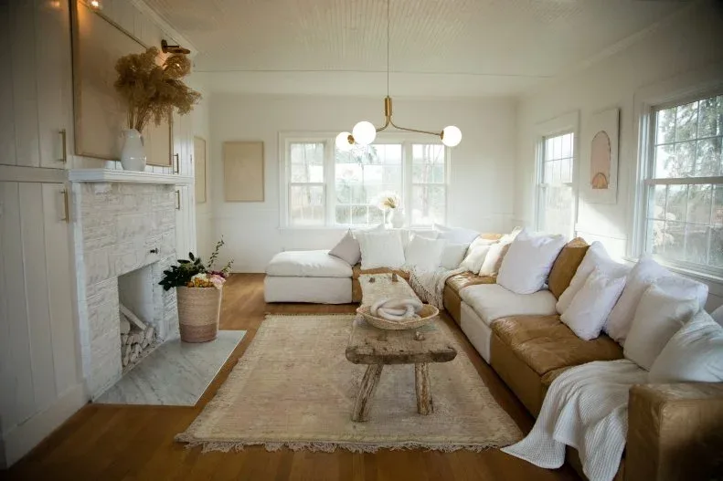 Light country room with white board walls, large tan-and-white sectional, rustic stool-shaped table and pale fireplace