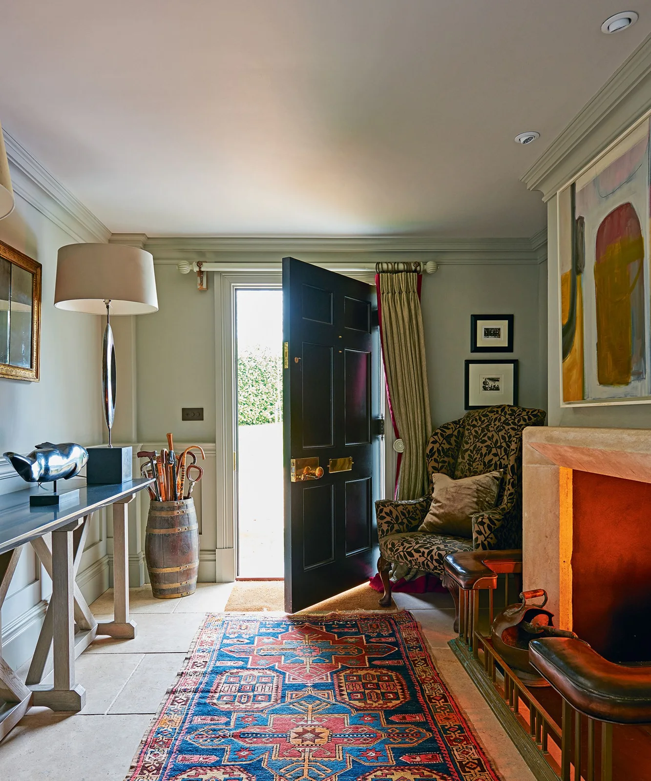 A patterned armchair sits beside a colorful rug, tall lamp, framed art and umbrella holder near a dark open front door