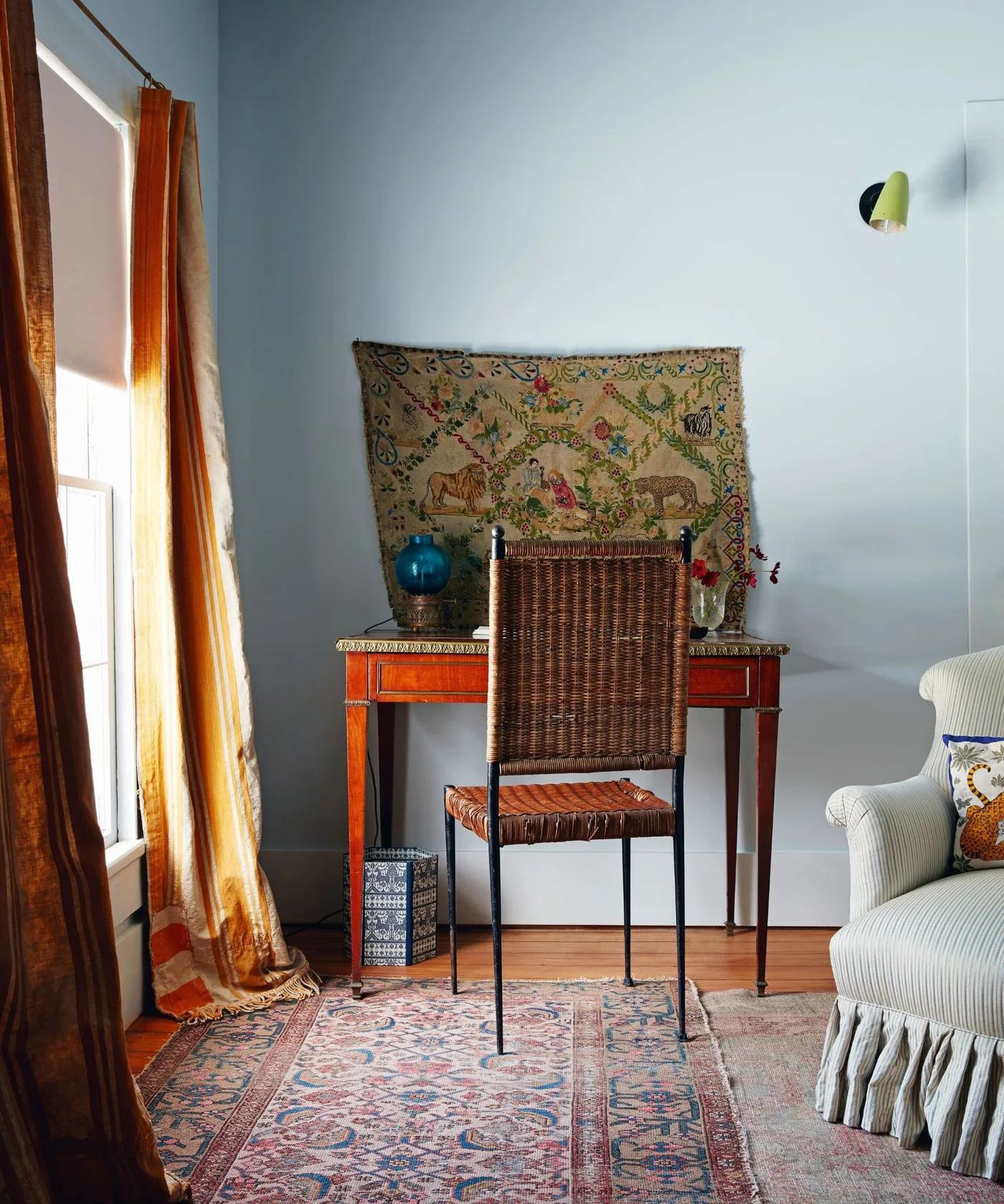 Bedroom corner with small wooden desk, woven high-backed chair, pale blue wall, orange curtains and patterned rug.