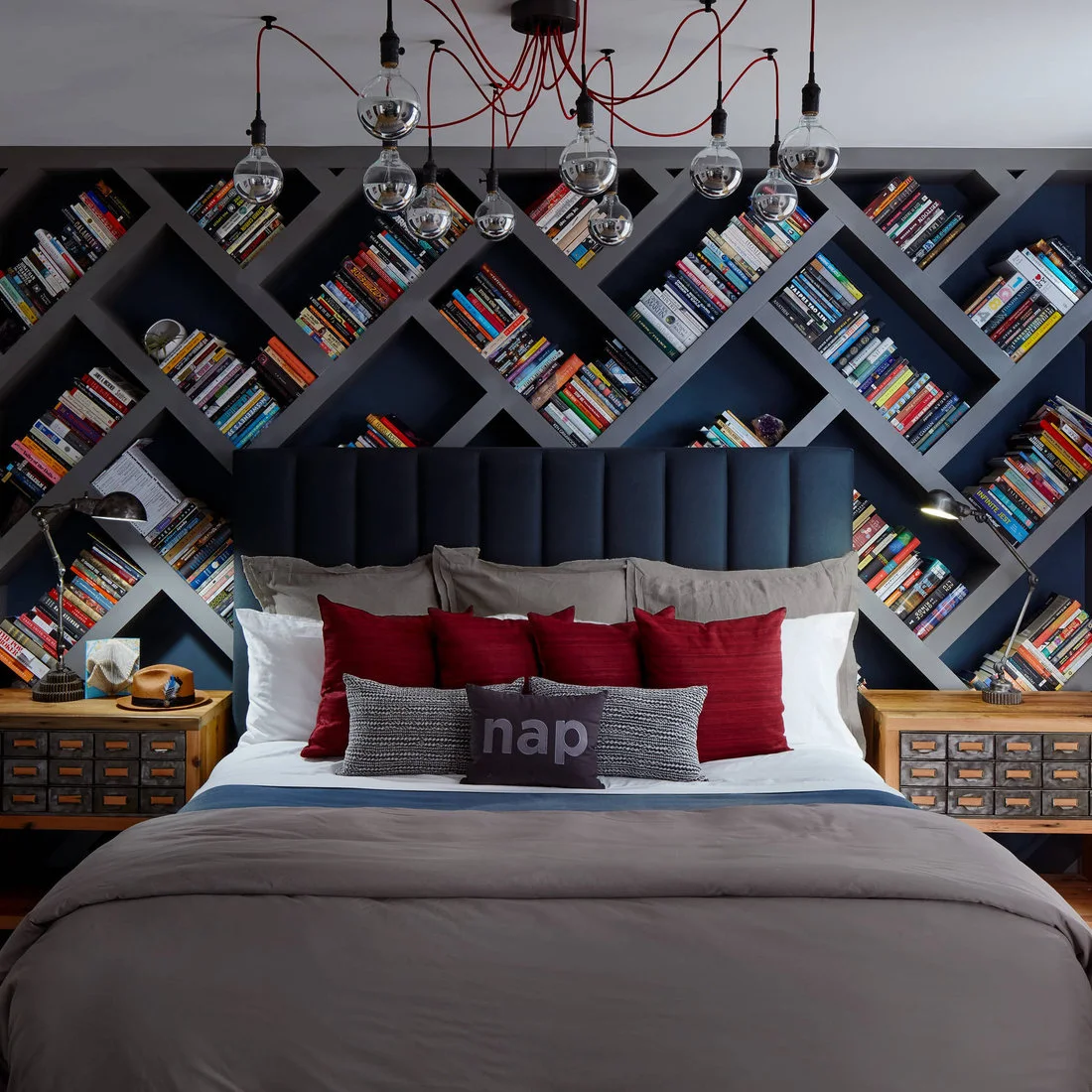 A navy upholstered headboard sits before diagonal dark bookshelves beside small wooden drawer cabinets and hanging bulbs on red cords