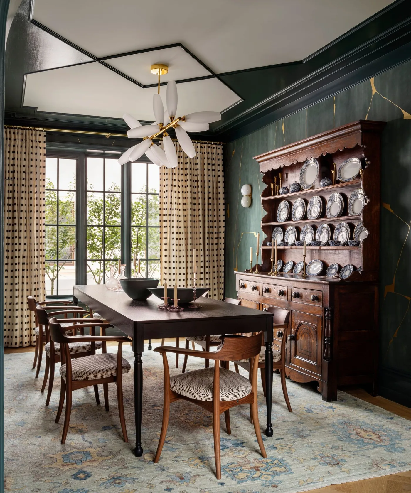 Dark green dining room with a wooden plate dresser, rectangular table, curved wooden chairs and patterned curtains.
