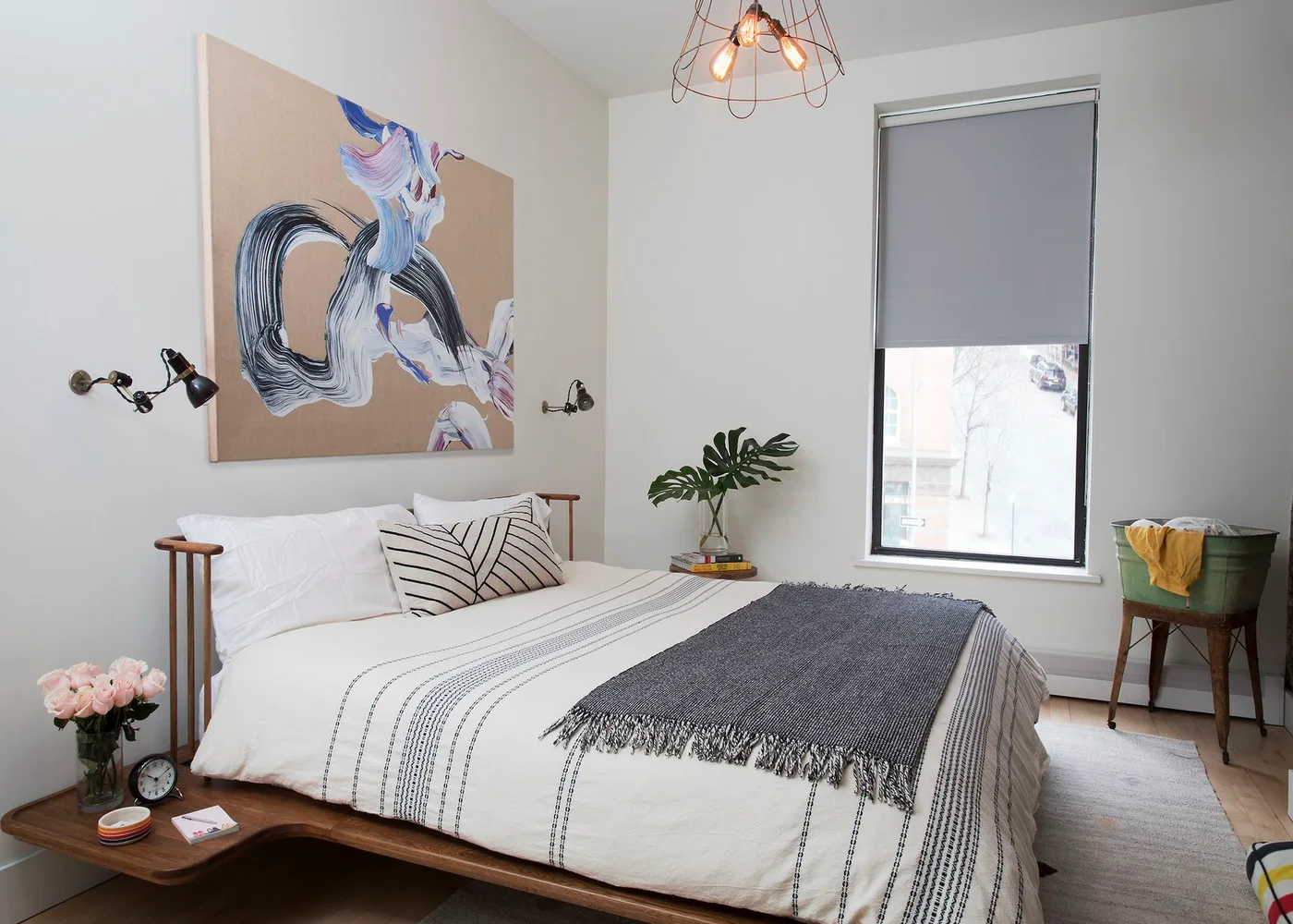 A low wood bedroom platform extends beside the mattress below a spindle headboard with wall lights, abstract artwork and pale patterned bedding