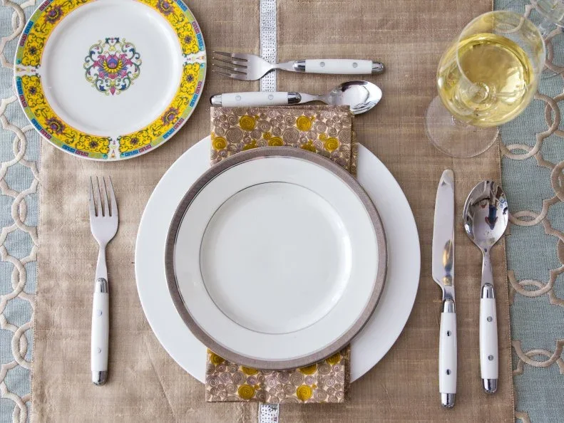 Plain white plates sit above patterned napkins beside a floral side plate, white-handled cutlery and clear glassware