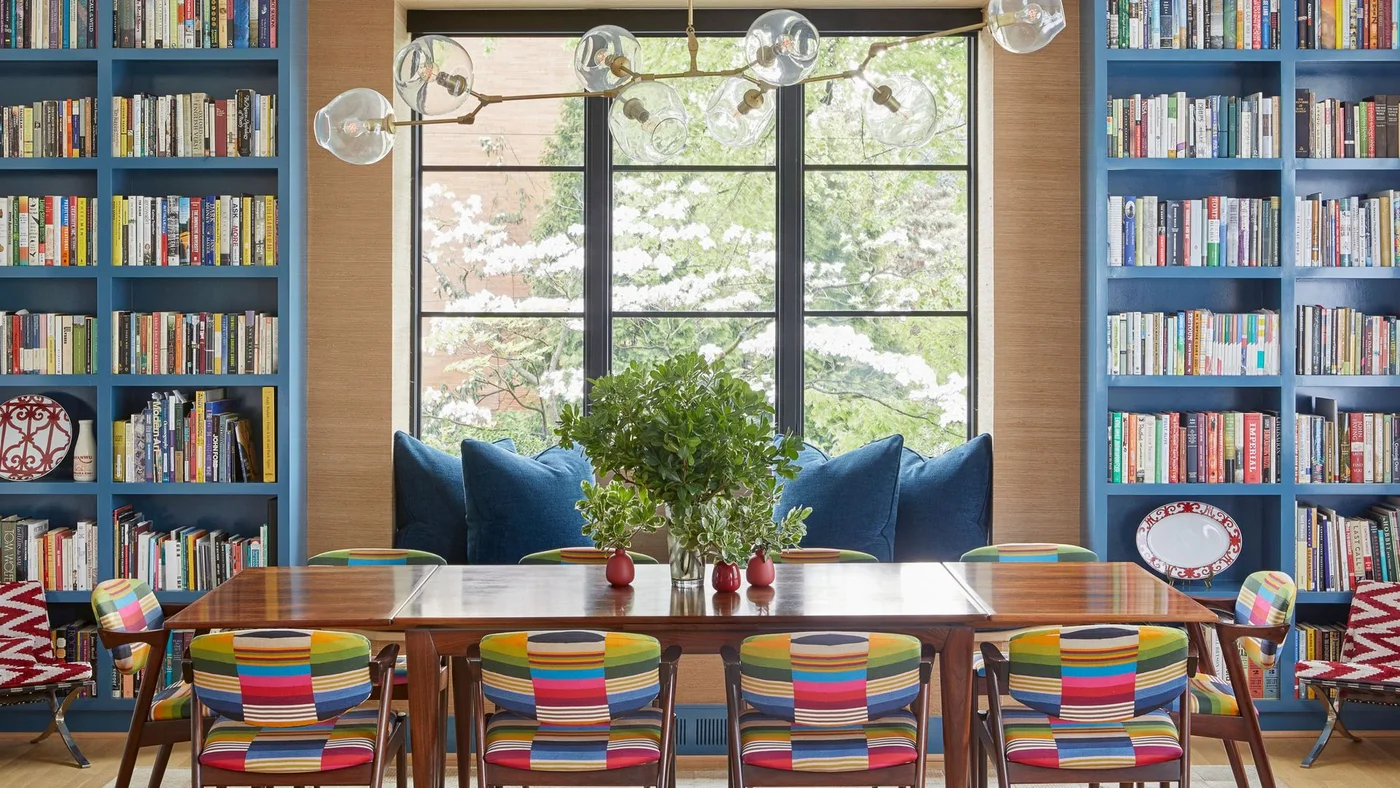 Colorful striped wood dining chairs surround a long wood table beside blue bookcases, a window seat and branching clear globe lights