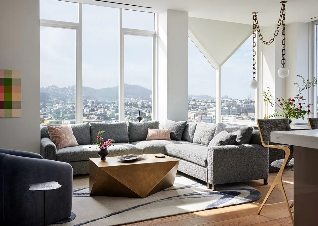 Bright living room with a gray corner sofa, faceted wooden coffee table, pale pink cushions and tall windows.