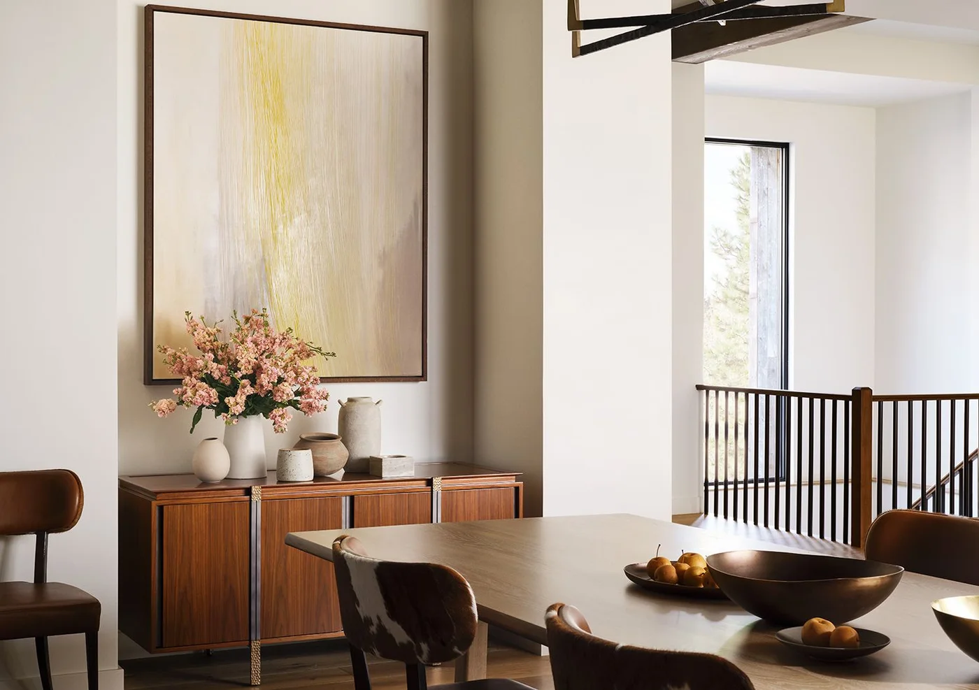 A warm wood sideboard stands beneath a large pale yellow abstract artwork beside a light dining table, curved brown chairs, pale walls and a dark stair railing