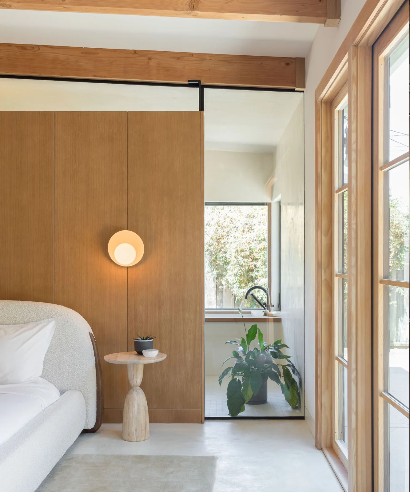 A pale curved upholstered bed sits beside a small wooden table and wall light against wooden panels near a tall glazed opening