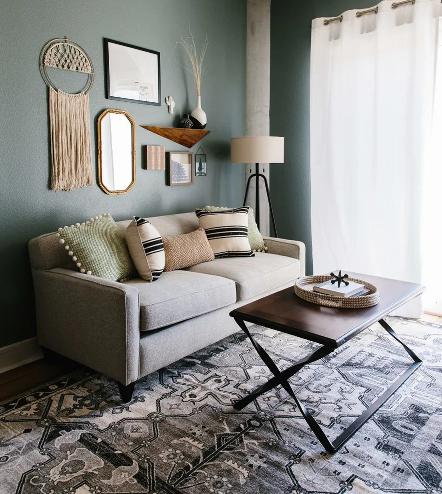 A framed picture, woven hanging, and mirror sit above a pale sofa on a muted green wall