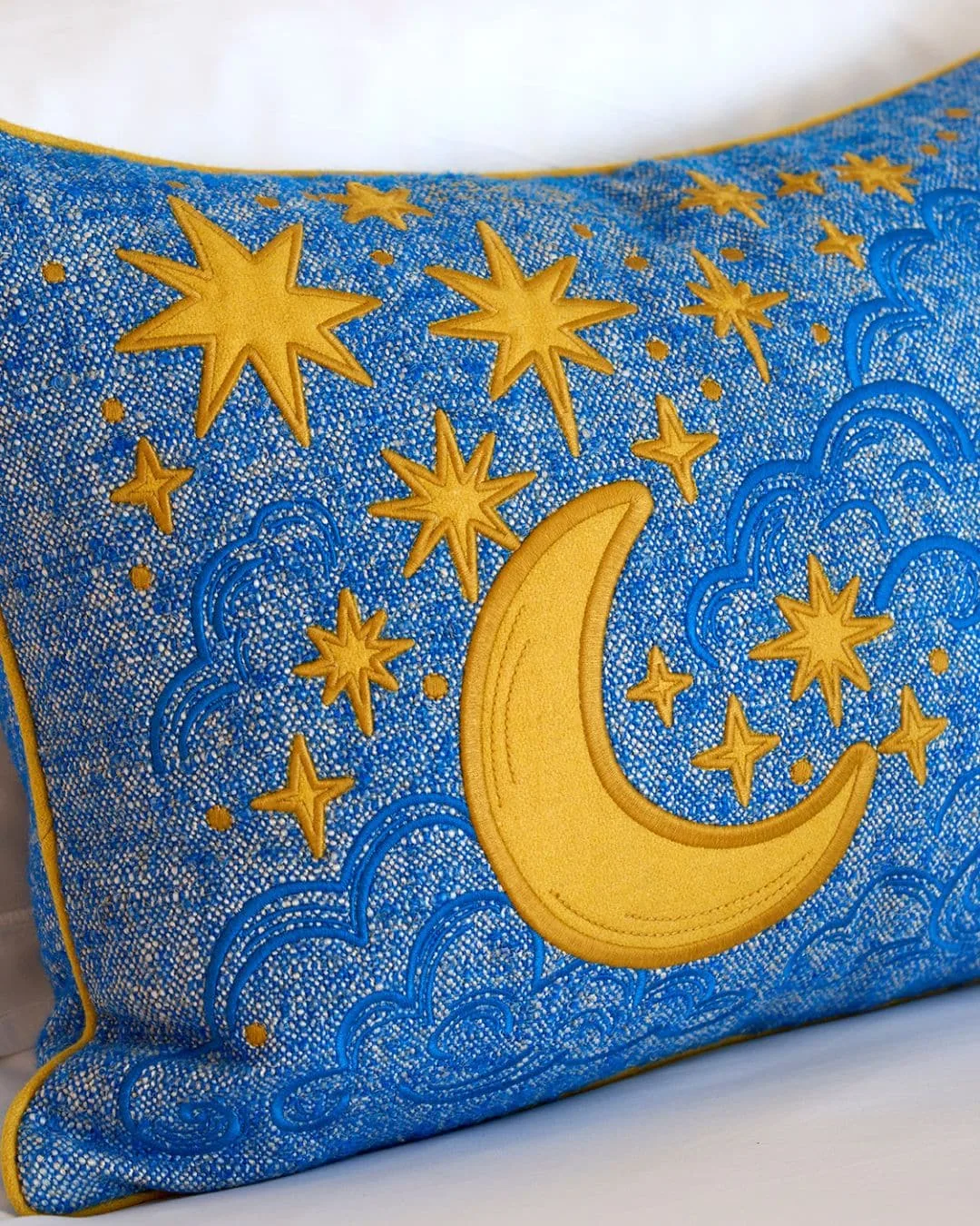 Blue decorative cushion with a yellow crescent moon, stars and swirling stitched patterns on white bedding.