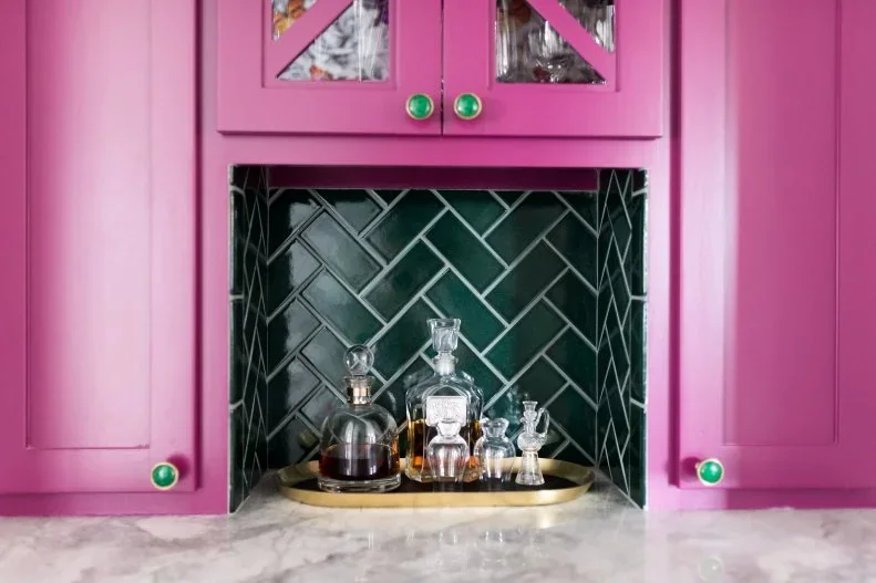 Deep green herringbone tiles line a small drink-storage nook within bright pink cupboards above a pale veined counter