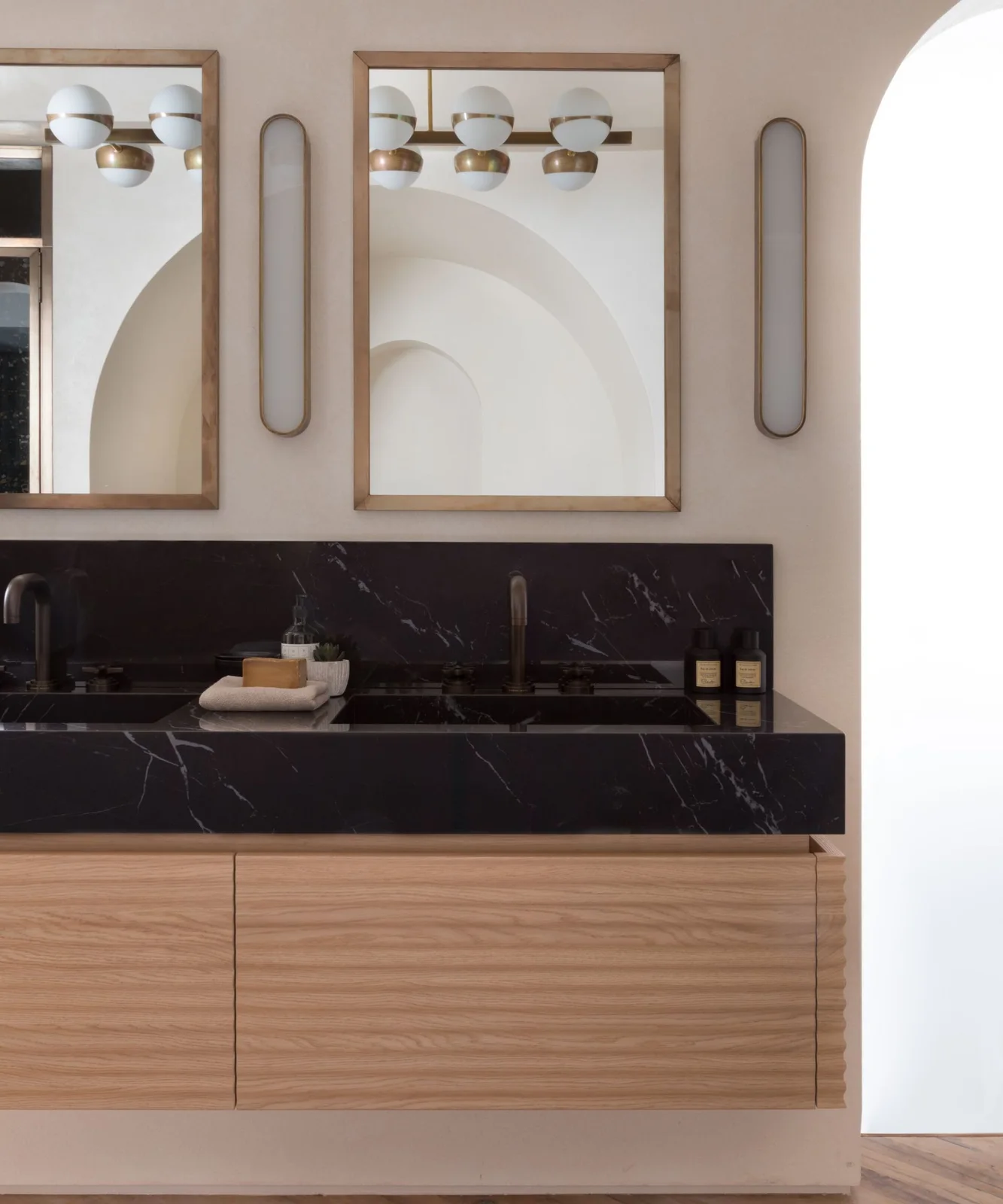 A wood double bathroom vanity has a boldly veined counter beneath two wood-framed mirrors and globe lights beside slim oval wall fittings