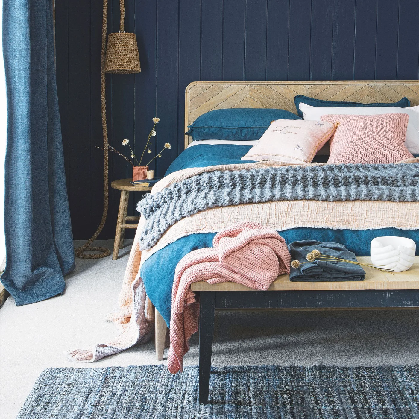 Bedroom with navy vertical paneling, pale wooden headboard, blue-and-blush bedding, woven pendant and a wooden bench.