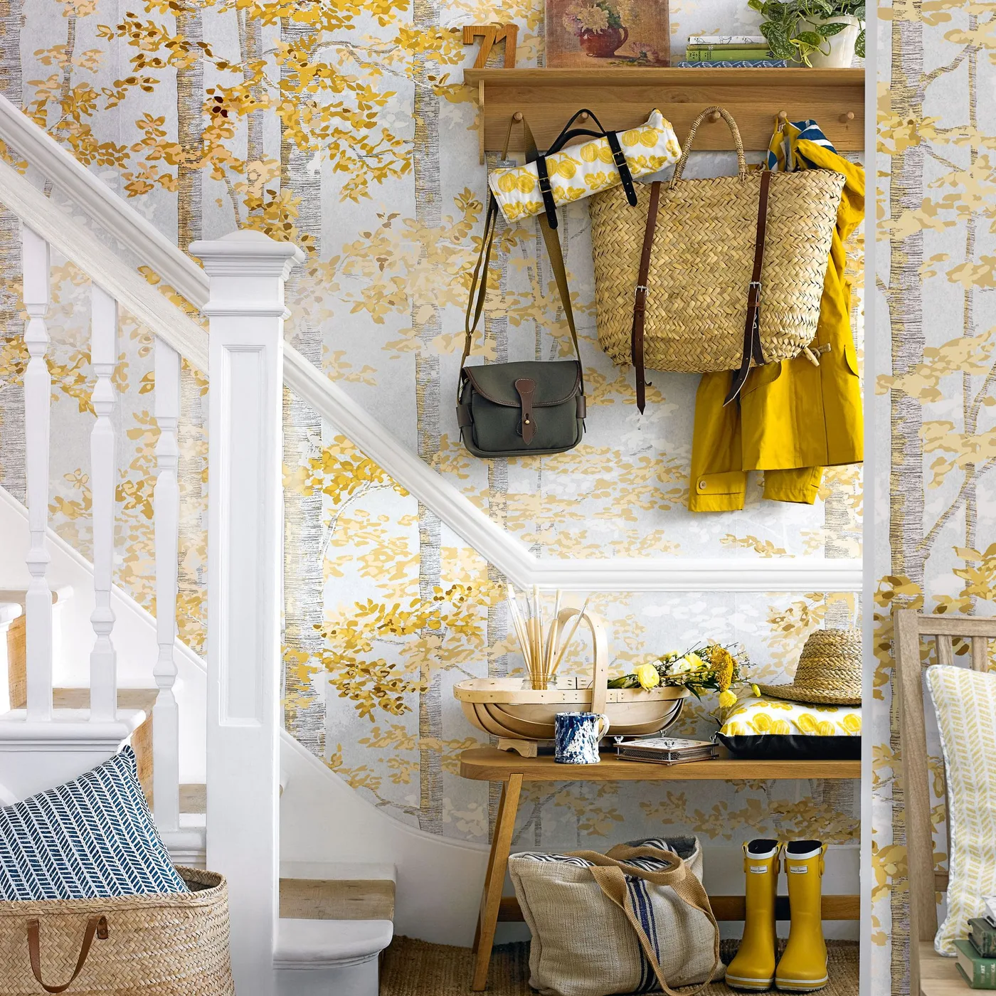 Country hallway with yellow leafy wallpaper, a wooden shelf and hooks, bags and a yellow coat, plus a small wooden bench beneath the staircase.