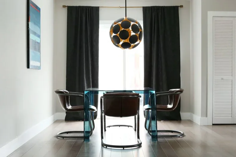 Dark dining armchairs with curved metal cantilever bases around a blue-edged glass table, with black curtains and a pale floor.