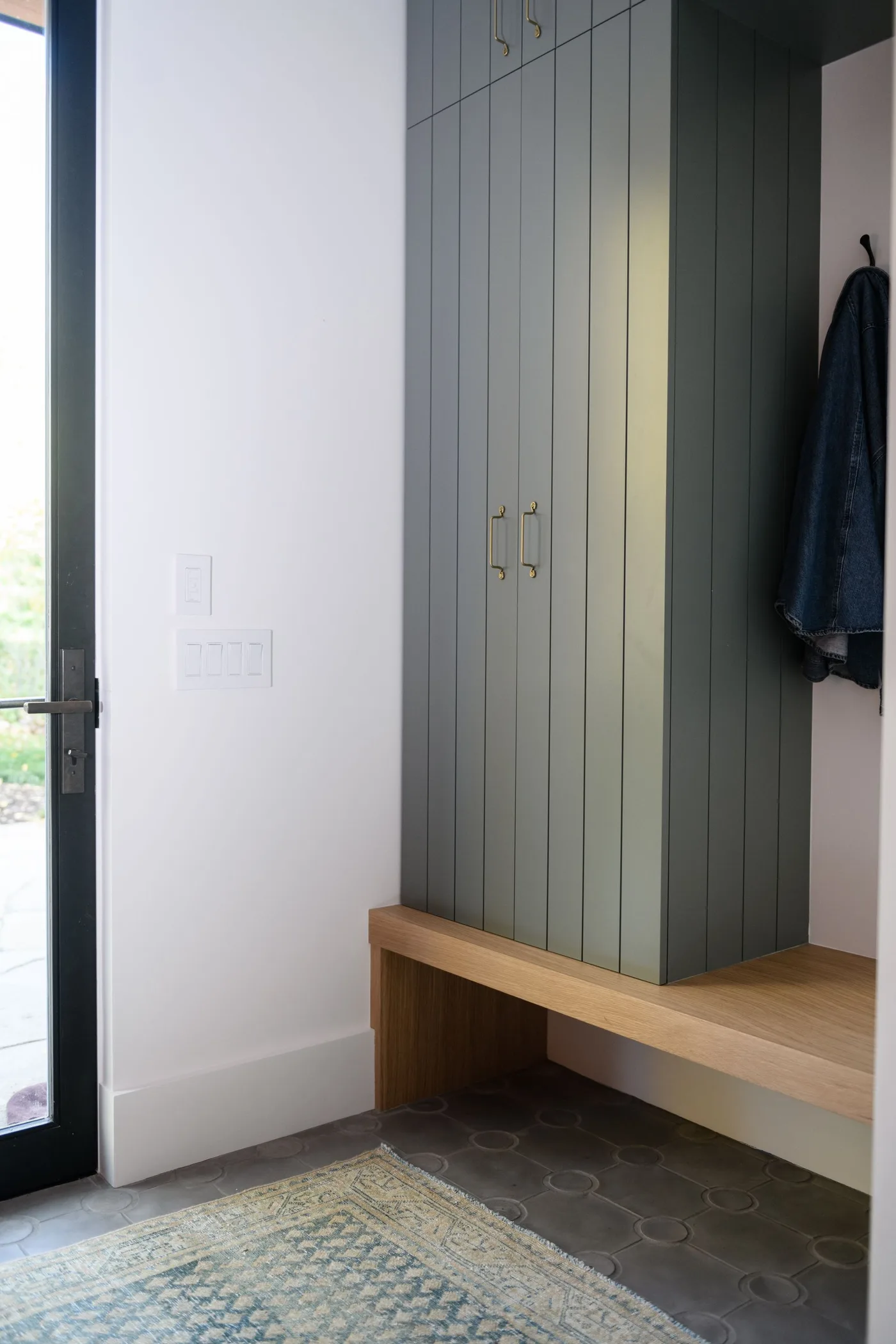 Small mudroom with muted green vertical cabinet doors, wooden bench turning along the side wall and dark floor.