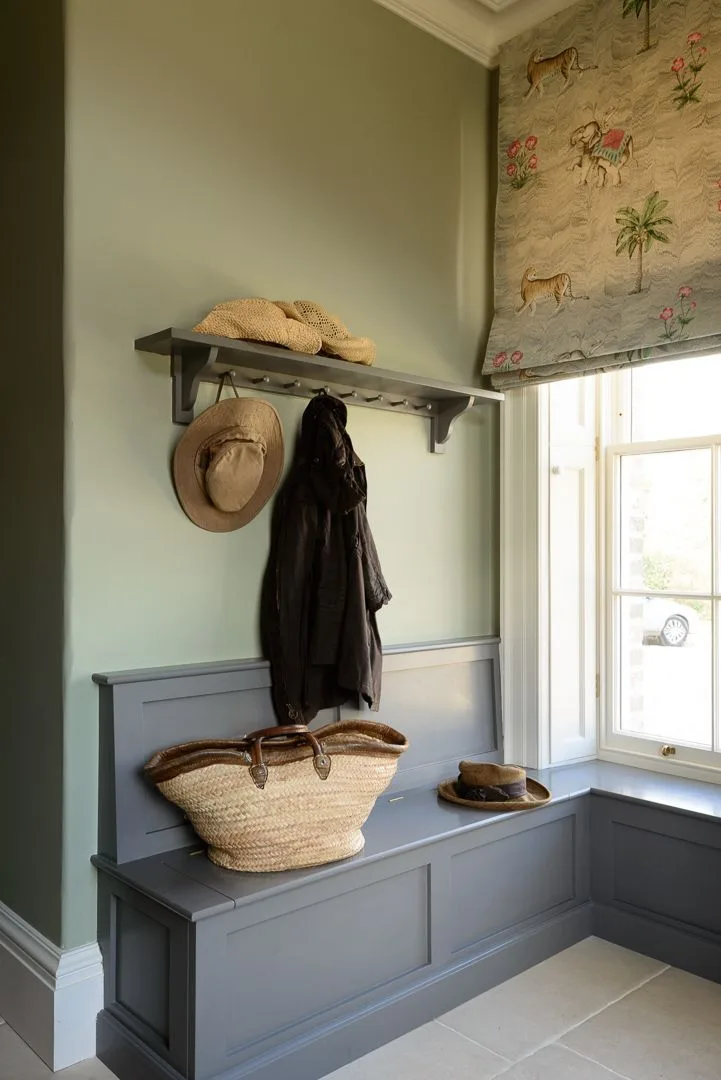 Gray corner bench beneath a hook shelf, with hats, coat and woven bag beside a window with a patterned blind