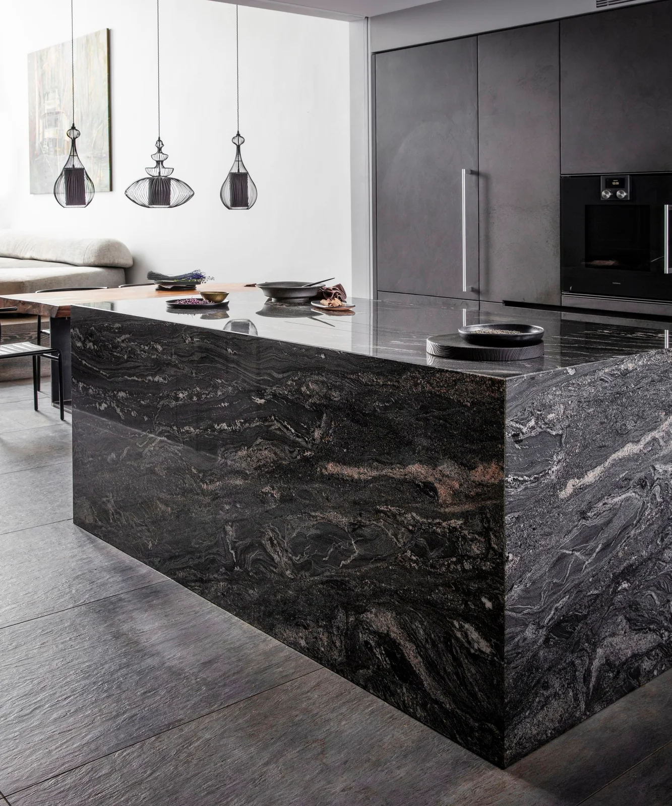 A strongly veined dark kitchen island stands beside flat gray tall cabinets below pale walls and three dark open pendant shades
