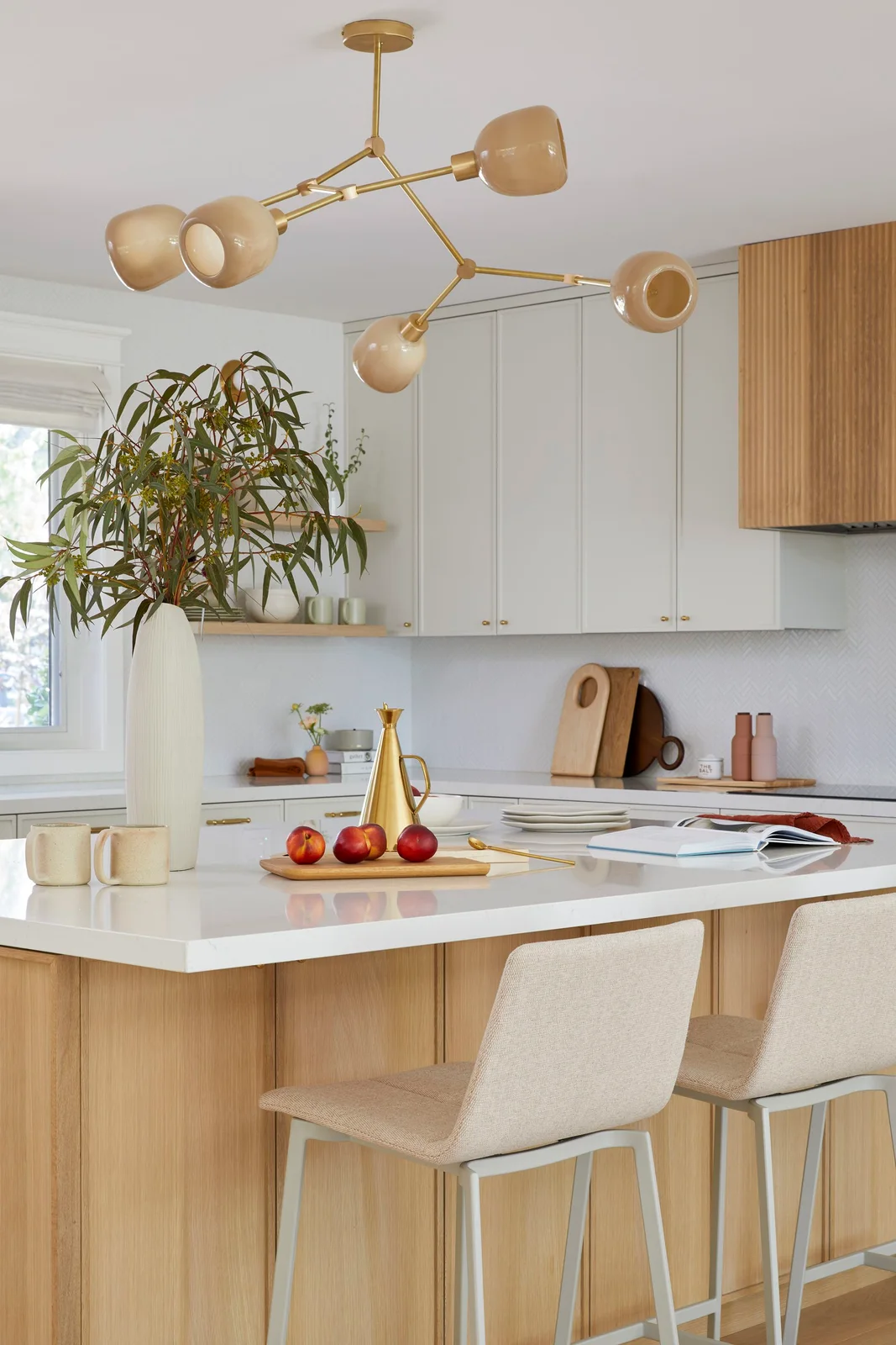 Pale kitchen cabinets and a wooden island with upholstered beige stools sit beneath warm-toned branching lights beside a wooden hood detail