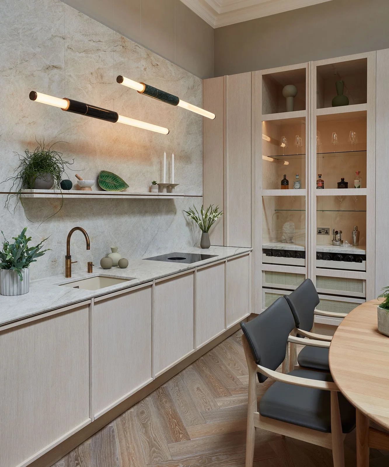 Pale wooden kitchen cabinets sit beside a glass-fronted drinks cabinet and long horizontal pendant lights