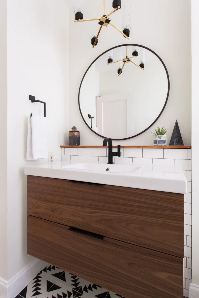 A floating wood vanity and white basin stand below a black round mirror beside black taps, white subway tiles and a narrow wood shelf