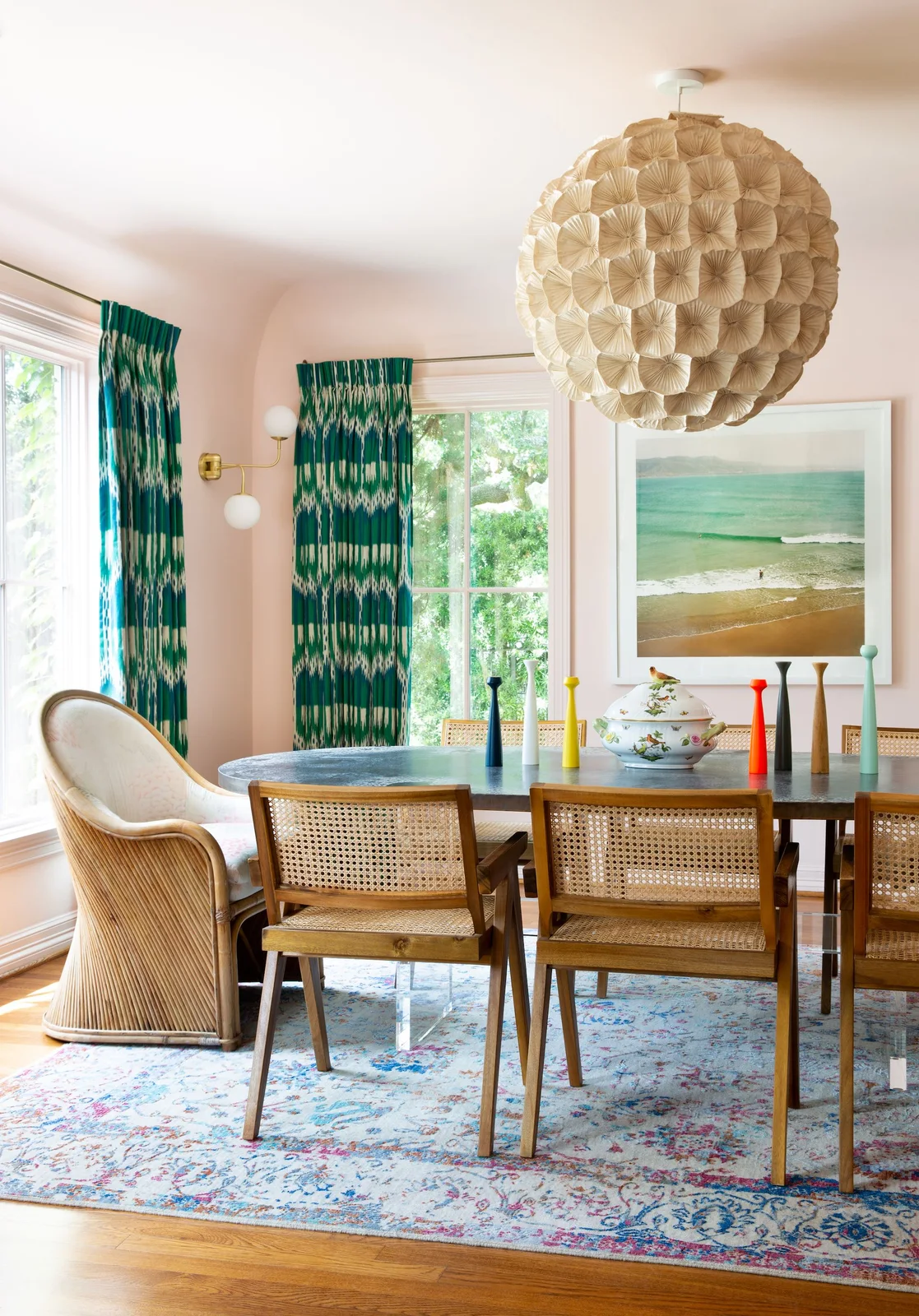 Pale pink walls surround green patterned curtains, a colorful rug, woven-back dining chairs and a large textured pale pendant