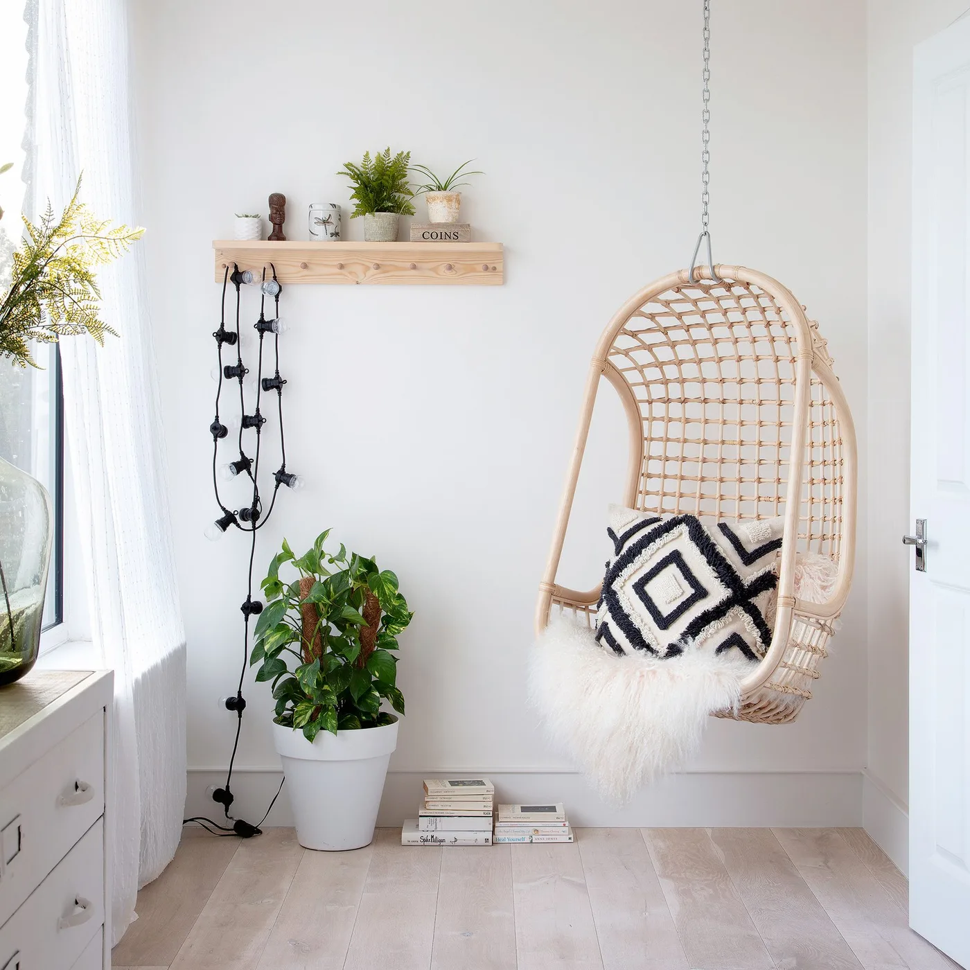 White bedroom corner with woven hanging chair, black-and-white pillow, floor plant, small plants on a shelf and pale drawers beside the window.