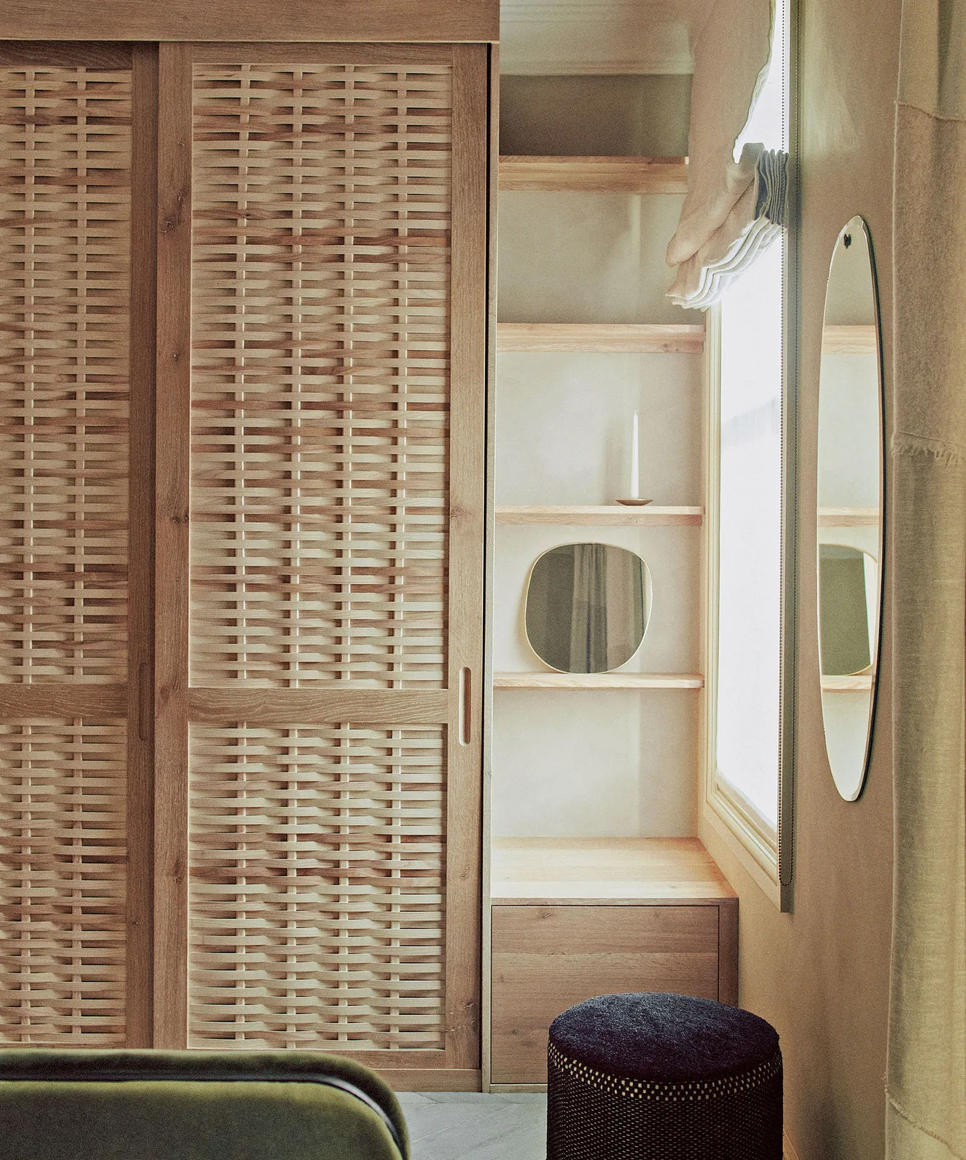 Bedroom storage corner with wooden woven-panel wardrobe doors, open side shelves, tall oval mirror and small upholstered stool.