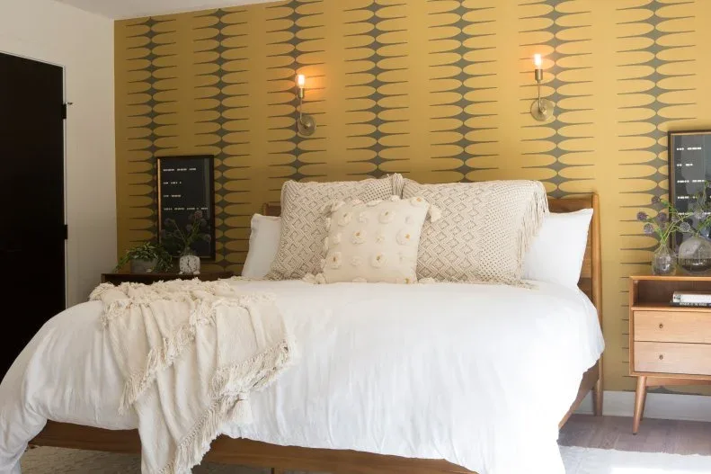 Ochre bedroom accent wall with repeated gray shapes behind pale bedding and a wood bedside unit