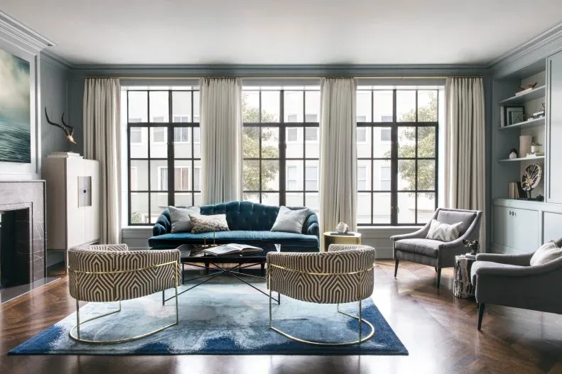 Living room with teal curved sofa, gray armchairs, two patterned chairs with rounded metal bases, glass table and blue rug in front of tall black-framed windows.