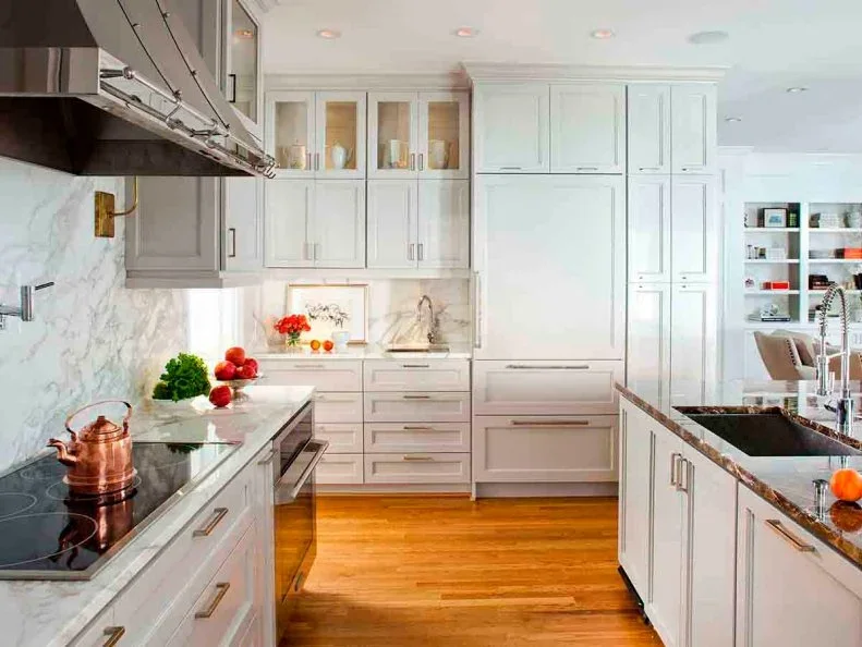 Pale framed kitchen cabinets and glass-front upper cupboards stand beside broad pale veined walls and counters with straight metal handles and warm flooring